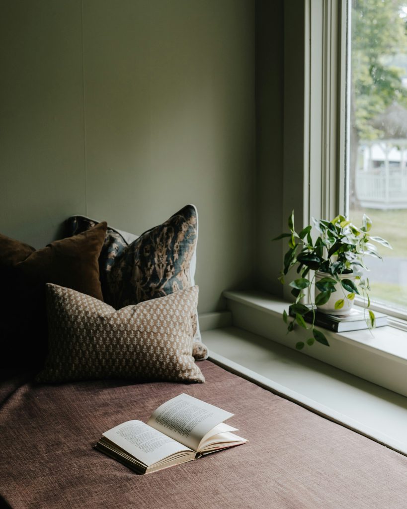 A calm, neutral living space with layered throw pillows, an open book on a window seat, and soft natural light creating a quiet, reflective atmosphere.