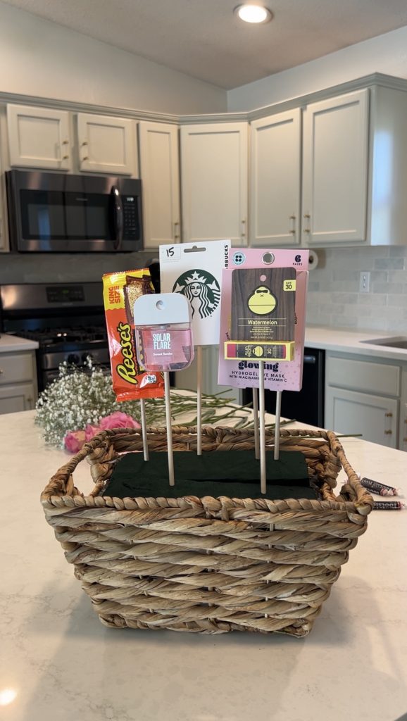 Gift basket items secured on wooden skewers inside a woven basket before adding flowers to complete the bloom basket arrangement.