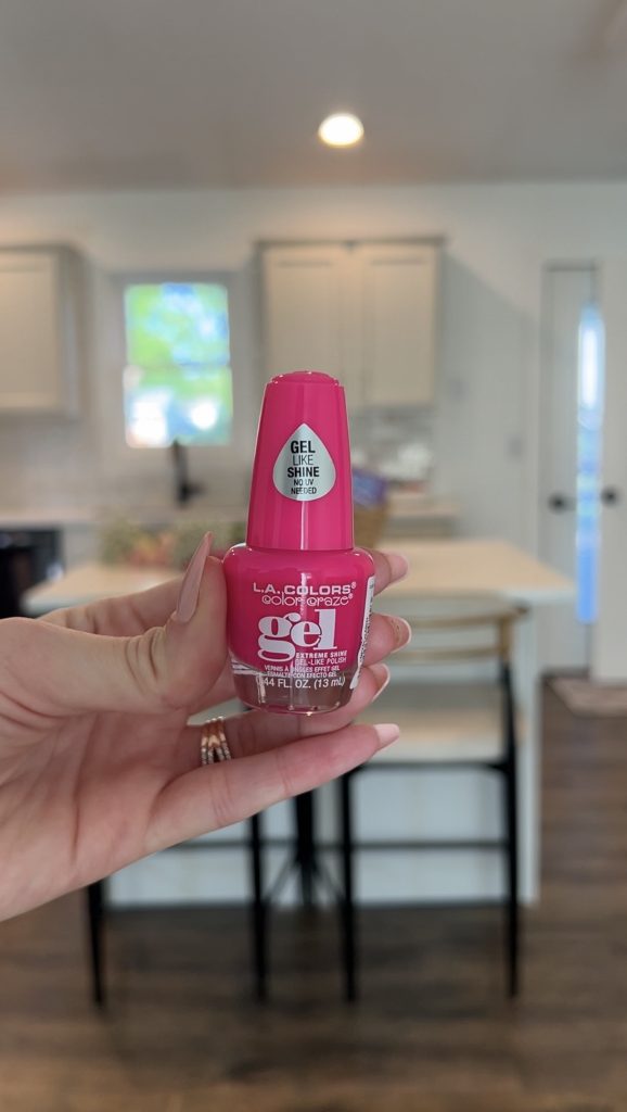 Hand holding bright pink gel nail polish in front of a kitchen background for a bloom basket gift idea.