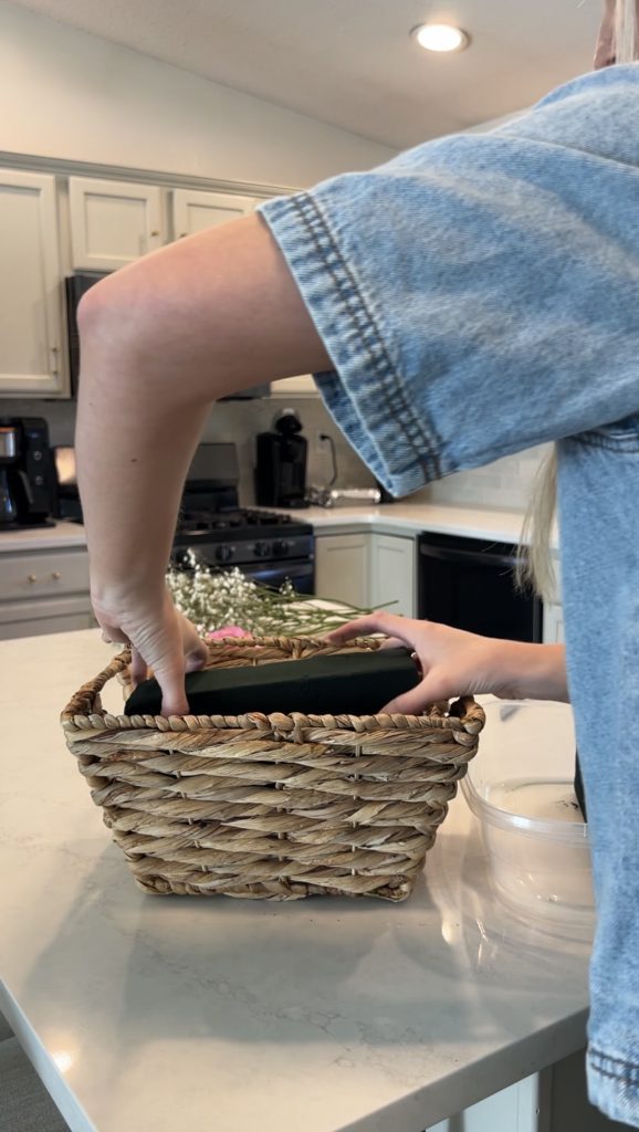 Placing soaked floral foam blocks into a woven wicker basket to create the base for a DIY bloom gift basket arrangement.