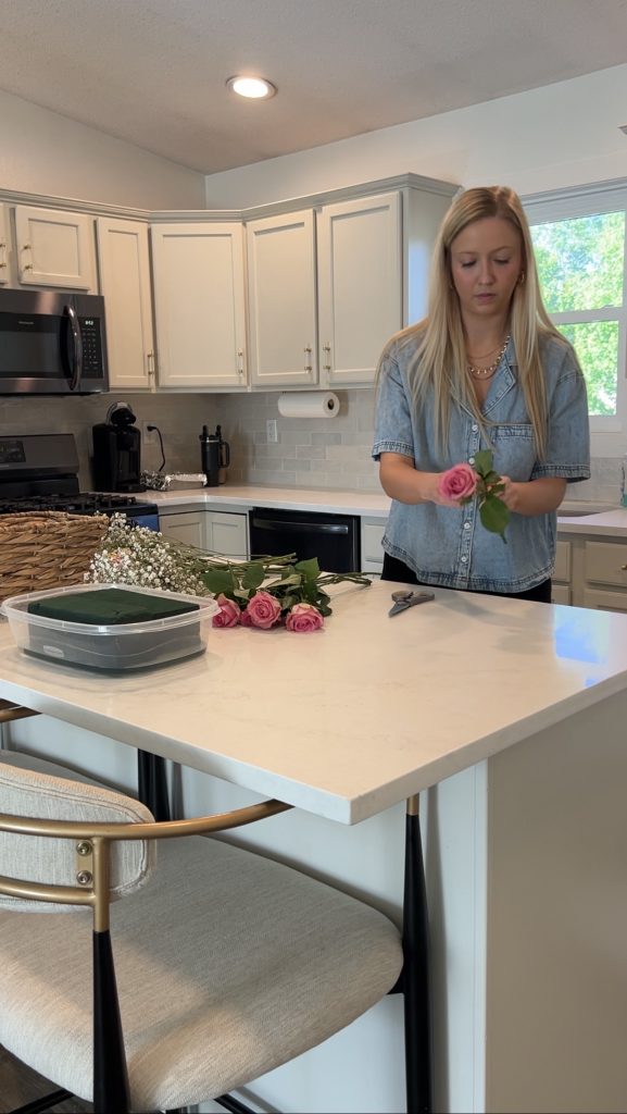Trimming and preparing pink roses beside floral foam and baby’s breath while assembling a DIY bloom basket on a kitchen island.