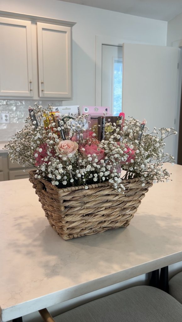 Finished DIY bloom basket arrangement featuring pink roses, baby’s breath, beauty products, candy, and a Starbucks gift card styled inside a woven wicker basket on a kitchen island.