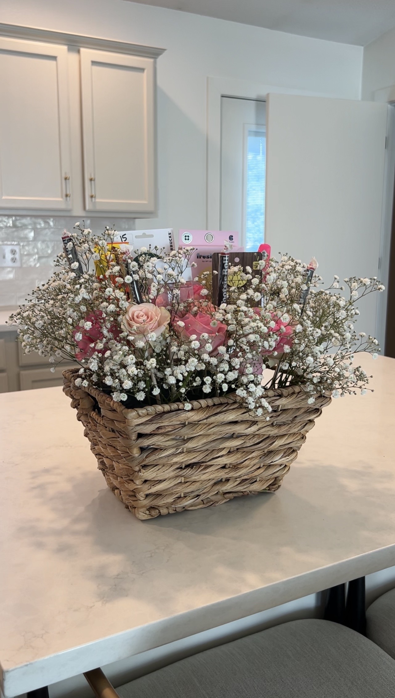 Finished DIY bloom basket arrangement featuring pink roses, baby’s breath, beauty products, candy, and a Starbucks gift card styled inside a woven wicker basket on a kitchen island.